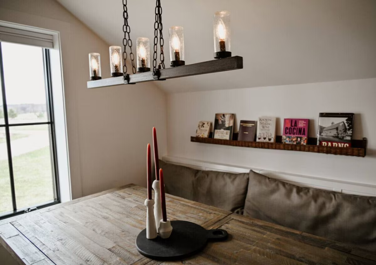 Dining area with a wooden table, a black candle holder with three tall candles, a window with a sliding glass door, a wall-mounted bookshelf with books, a sofa with cushions, and a rustic chandelier with six glass-encased light bulbs hanging from cha