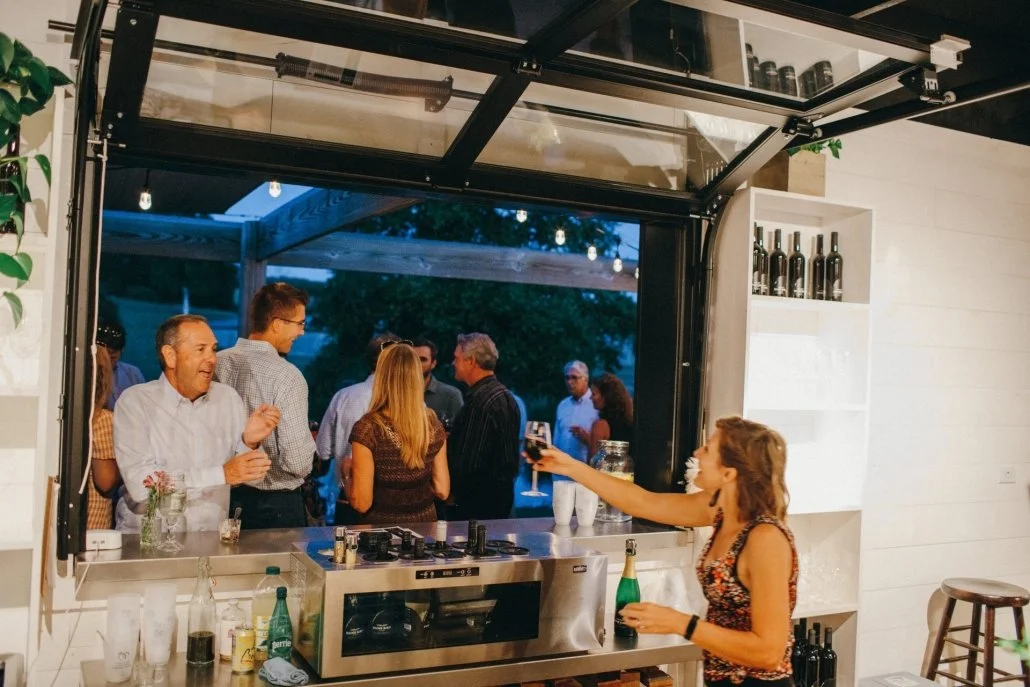 A woman inside a kitchen serving drinks to a group of people at an outdoor gathering.