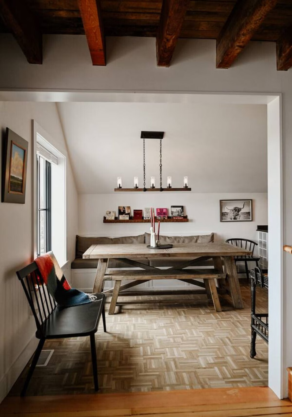 A cozy dining room with a wooden table, bench seating, and black chairs. Artwork and books decorate the white walls, with a window letting in natural light. A wrought iron chandelier hangs above the table, and a red and black throw is draped over one