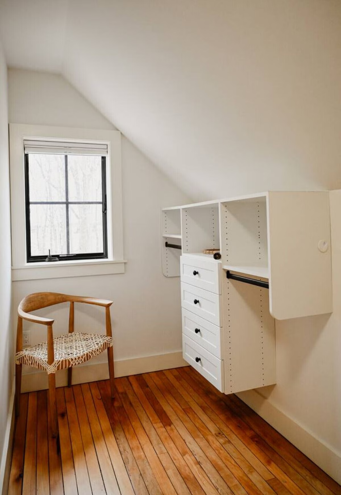 A small room with a window, a wooden chair with a woven seat, and white shelving with drawers and hanging rods.