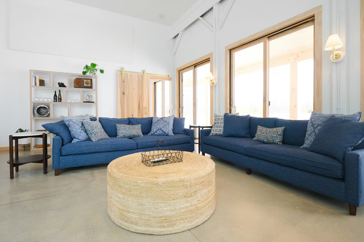 Living room with two blue sofas, a round woven coffee table, a white bookshelf, and large windows letting in natural light.