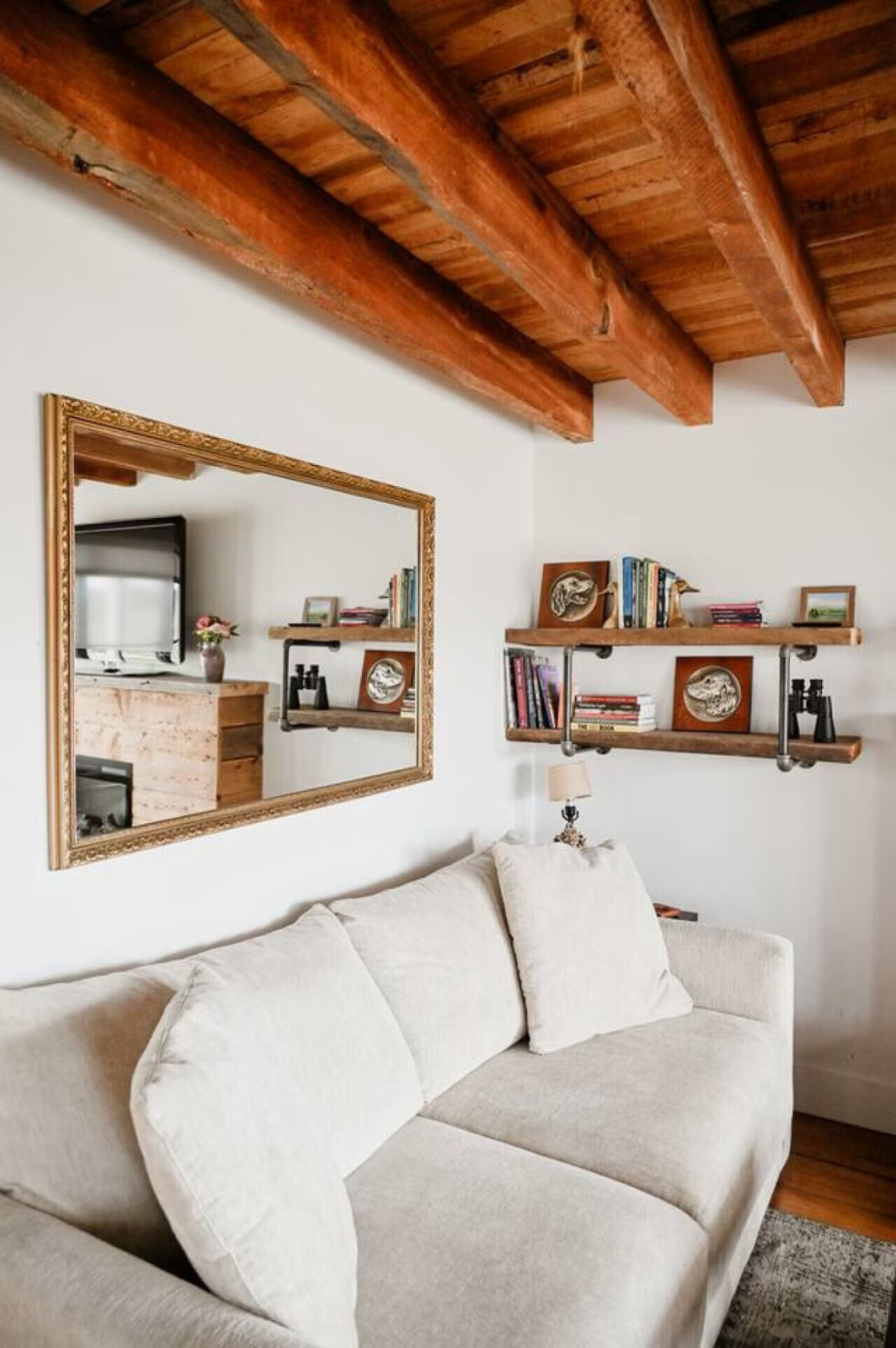 Living room with a white sofa, a large mirror on the wall, wooden ceiling beams, and two wooden shelves with books and decorative items.