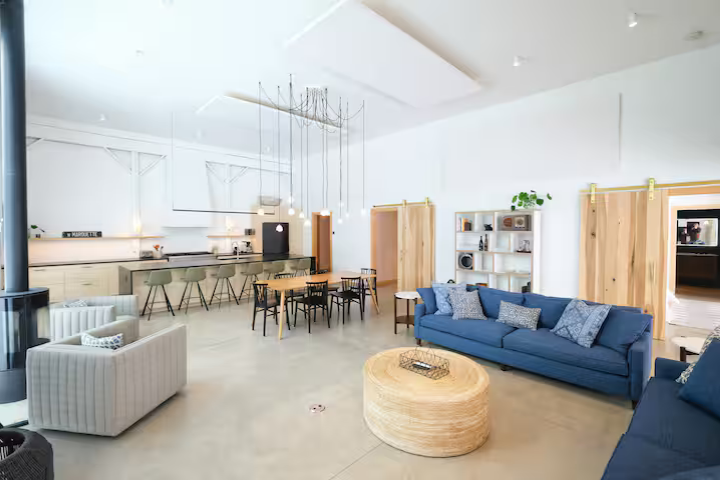 A spacious, modern open-plan living area with a kitchen, dining area, and seating. The room features white walls, high ceilings, a blue sofa, a round wooden coffee table, and a white armchair. There are wooden sliding doors, shelves with decor, and h