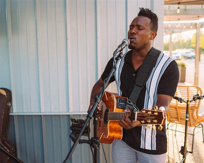 A man playing an acoustic guitar and singing into a microphone outdoors.