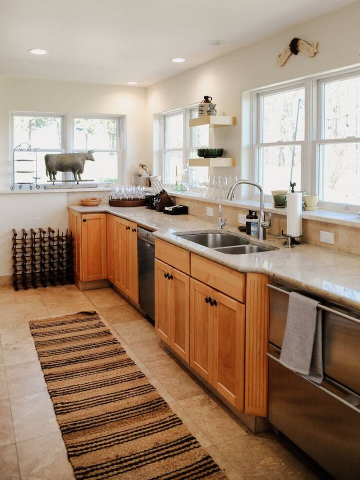 A kitchen with wooden cabinets, a double sink, and large windows allowing natural light. The countertop has wine glasses, a paper towel holder, and decorative items. There is a striped rug on the tiled floor and wall-mounted shelves with plants and d