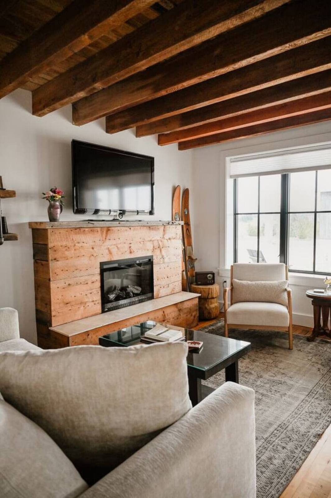 A cozy living room with a beige sofa, a black coffee table with books, a beige armchair, and a large window with a view outside. There is a fireplace made of wooden planks, a flat-screen TV above it, and wooden beams on the ceiling. Decor includes a 
