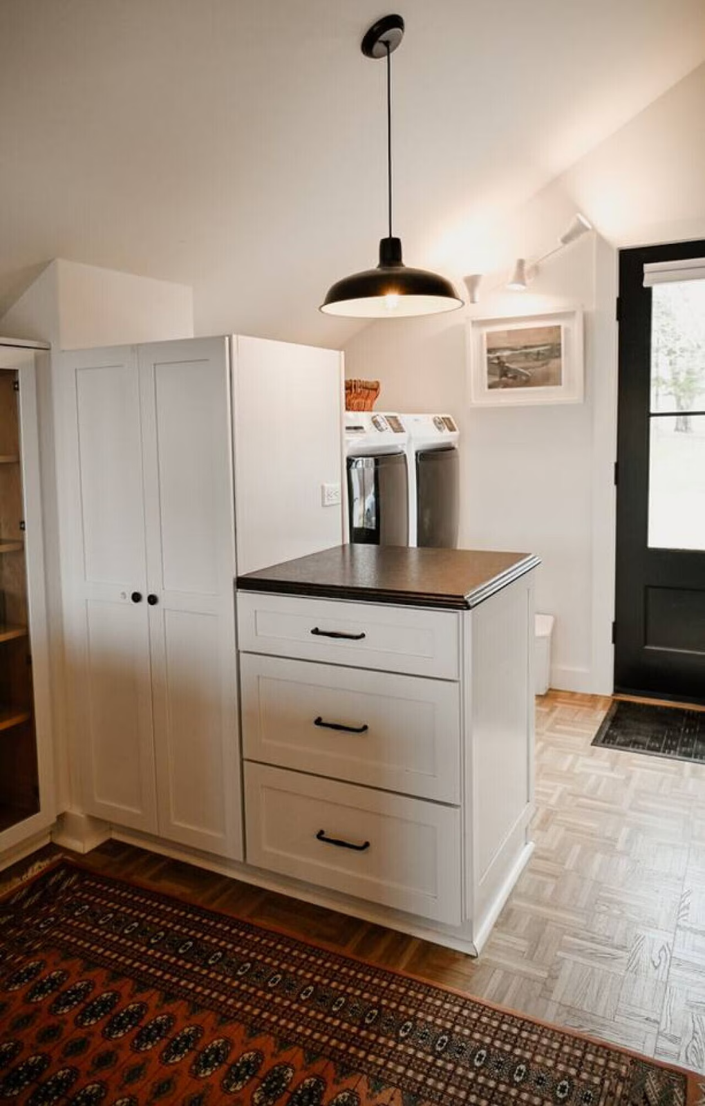 A laundry room with a white cabinet, a mini island with black handles, a washer and dryer, a black ceiling light, a picture on the wall, a black door, and a patterned area rug.