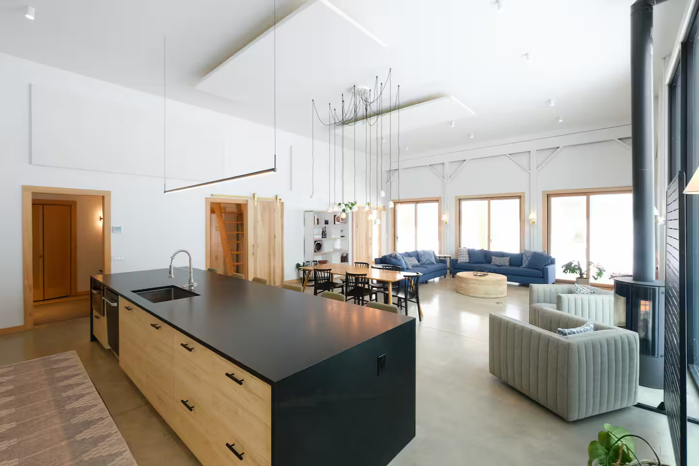 Open-concept living and dining area with a kitchen island, a blue sofa, a dining table with black chairs, armchairs, and large windows in a modern home.