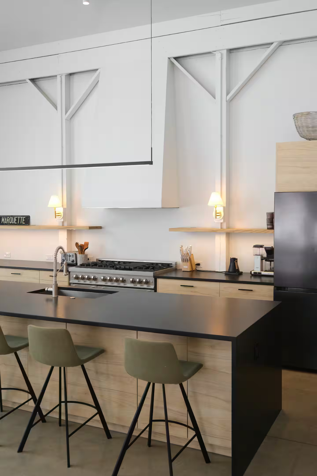 Modern kitchen with black island, beige cabinets, stainless steel stove, and minimalist decor, including wall-mounted lights and small appliances.