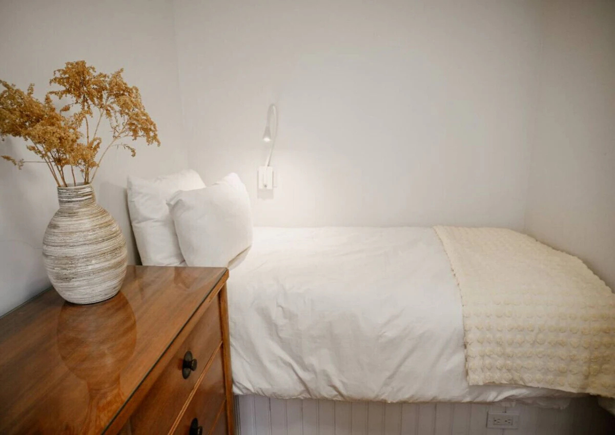 A simple bedroom with a bed, white bedding, pillows, and a blanket. There is a wooden nightstand with a large vase of dried flowers and a wall-mounted reading light.