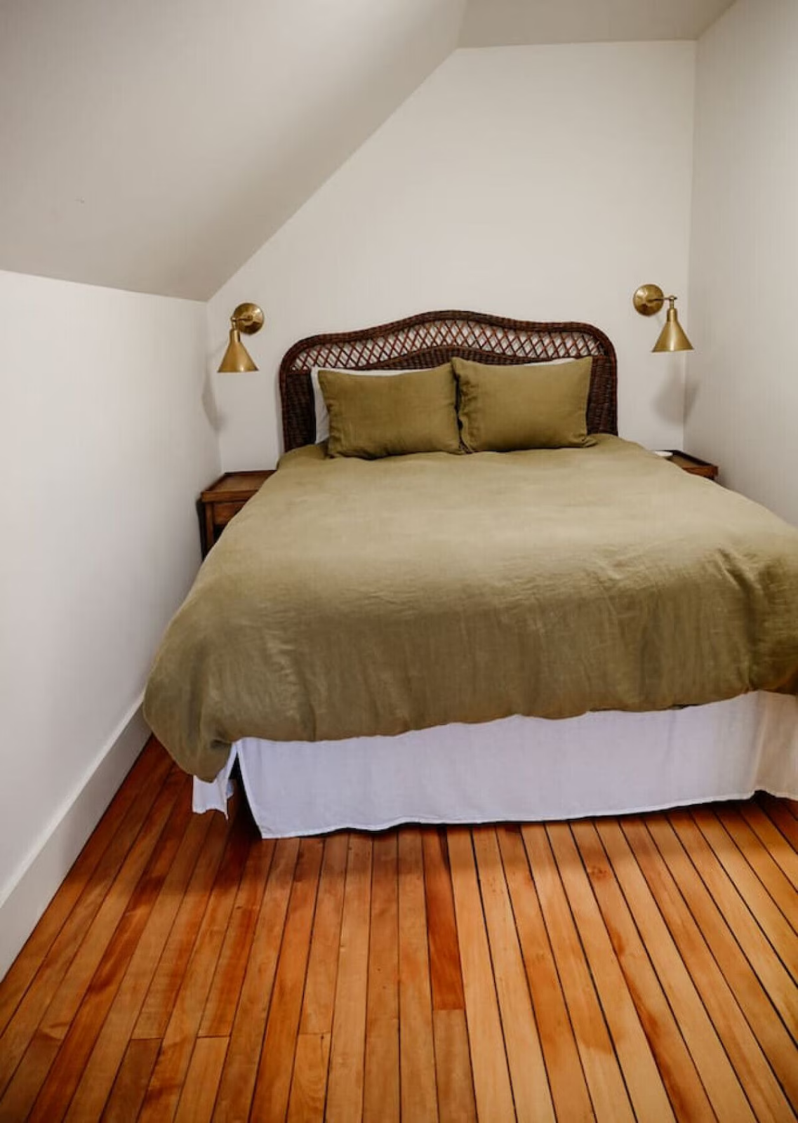A neatly made bed with an olive green bedspread and matching pillows, set against a white wall with sloped ceiling. Two wall-mounted brass reading lights are above the bed, with wooden nightstands on either side. The room has a hardwood floor.