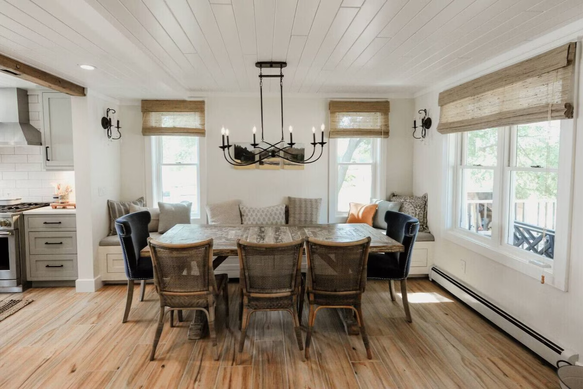 A dining room with a large rustic wooden table, six chairs, built-in window seat cushions, large windows with bamboo blinds, a black chandelier, and a view of a portable stove and kitchen area.