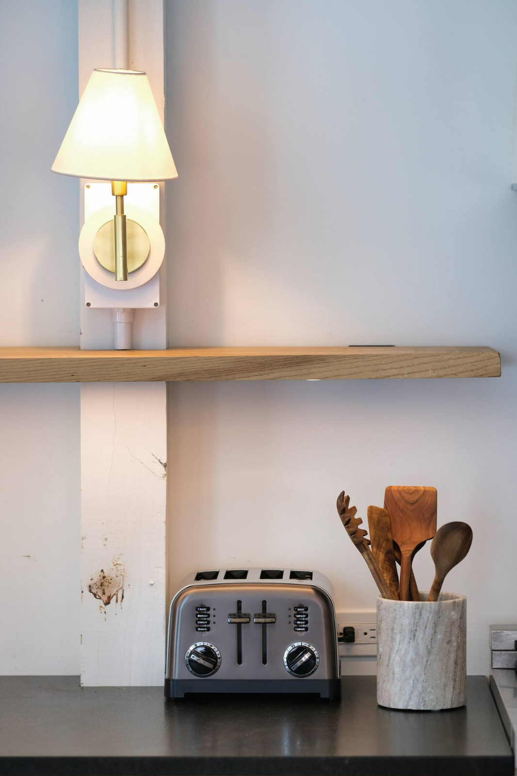 A modern kitchen countertop with a stainless steel toaster and a beige marble utensil holder filled with wooden utensils, and a wall-mounted lamp above the counter.