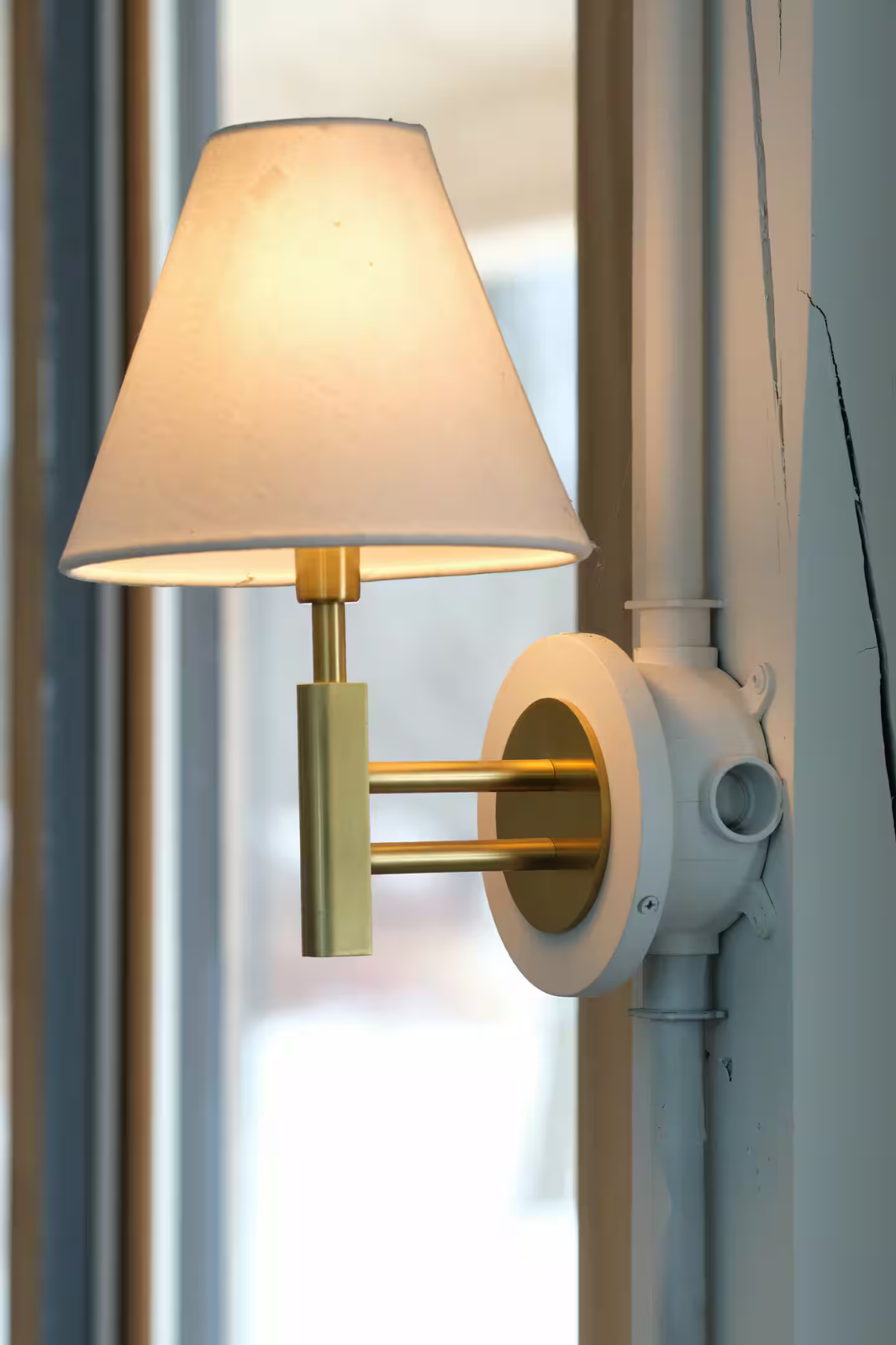 A wall-mounted lamp with a beige lampshade and a brass fixture, attached to an electrical socket on a wall.
