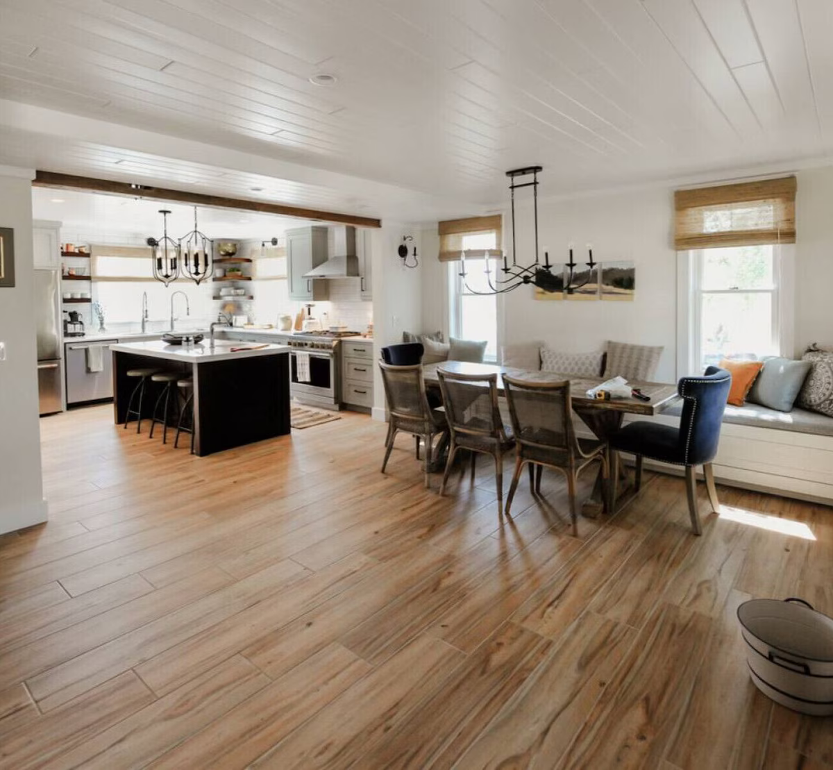Open concept kitchen and dining area with light wood flooring, white cabinets, kitchen island, hanging light fixtures, dining table with chairs, window seat, and natural light coming through windows.
