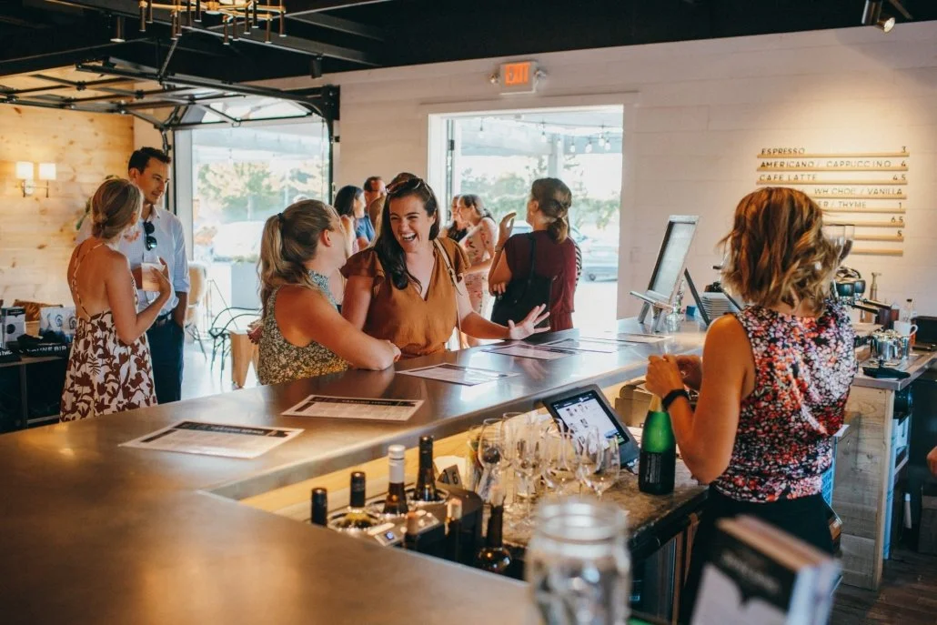 People ordering and chatting at a modern cafe counter with a menu on the wall and bottles of alcohol on the counter.