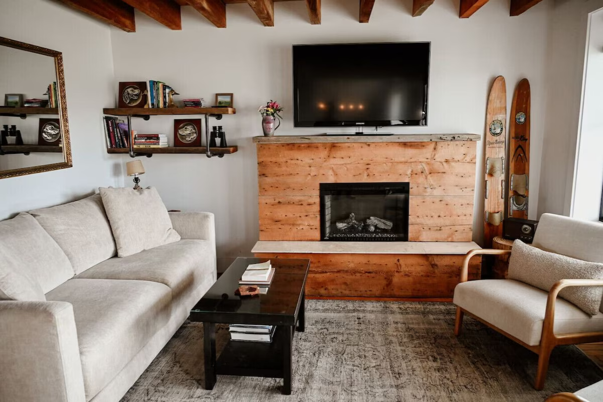 Living room with beige sofa and armchair, black coffee table, wooden fireplace mantel, flat-screen TV, decorative skis, wall mirror, and shelves with books and decor.