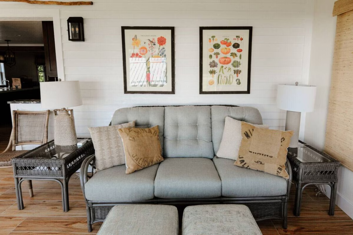 Living room with a gray sofa, patterned throw pillows, and two wicker side tables with lamps. Art prints hang above the sofa, and a wicker chair is visible on the left. The wall is white with horizontal plank paneling.