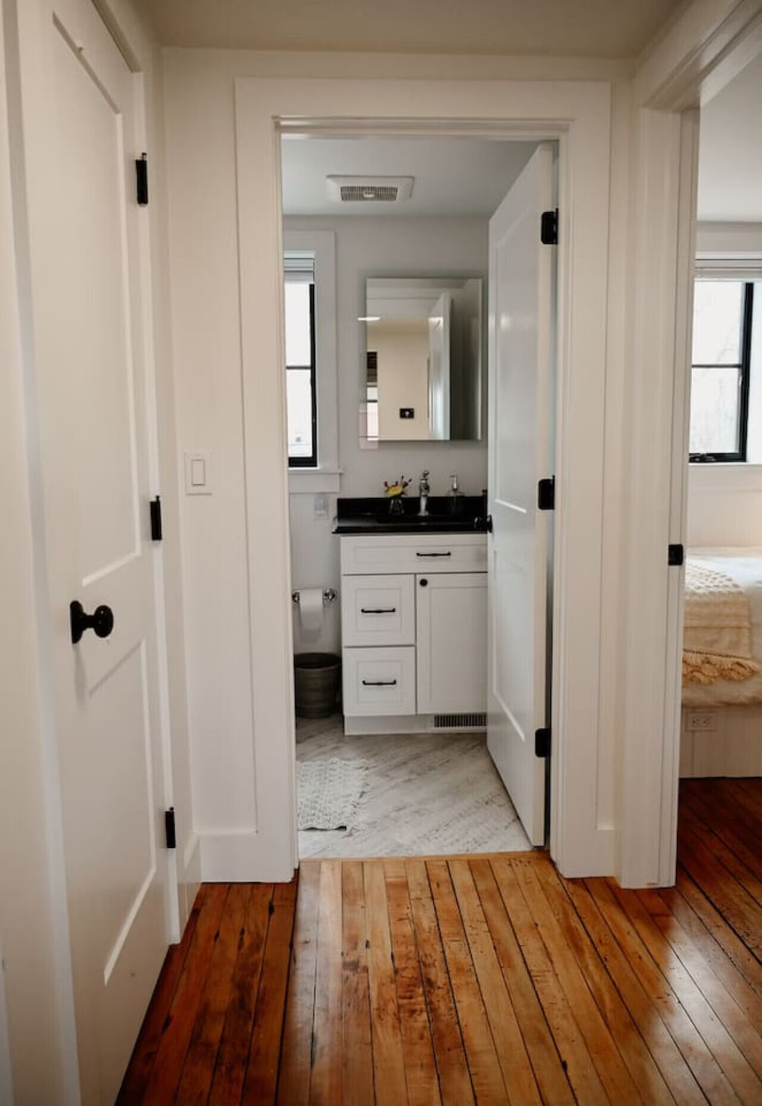 A view into a small bathroom from a hallway, featuring a white vanity with a mirror, two sinks, and black hardware, with a window and part of a bedroom visible.