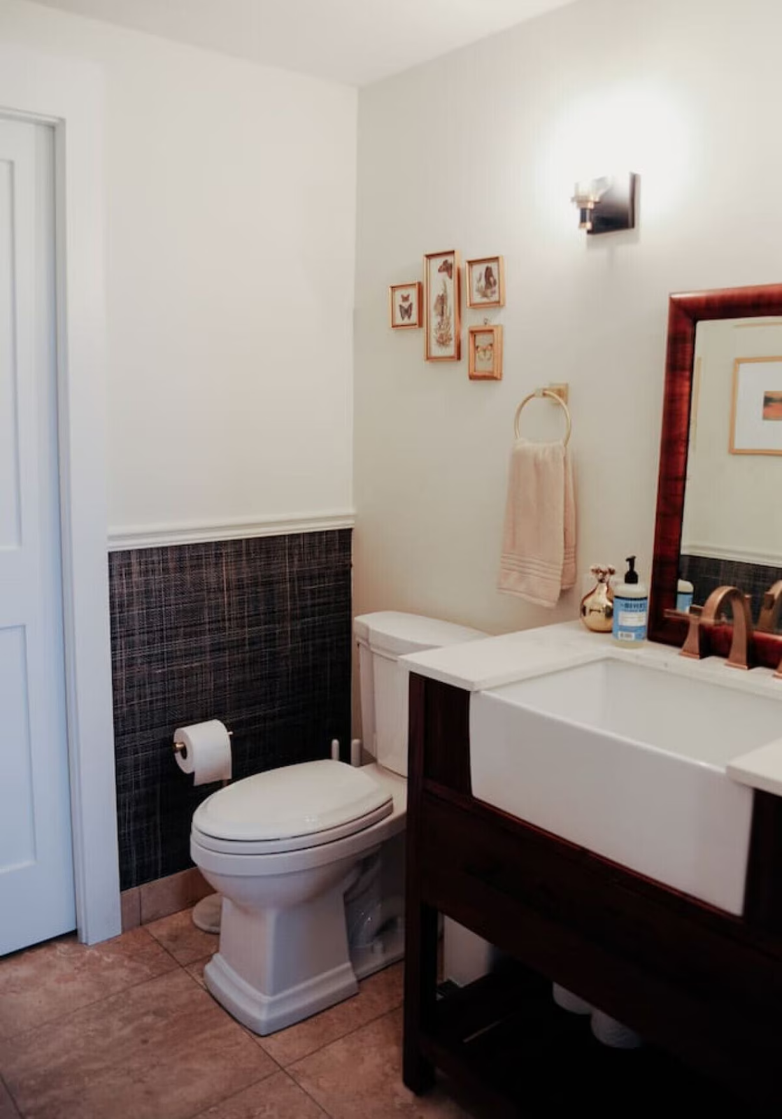 A small bathroom with a white toilet, a dark wood vanity with a white sink, a wall mirror, and framed butterfly and flower pictures on a cream-colored wall.