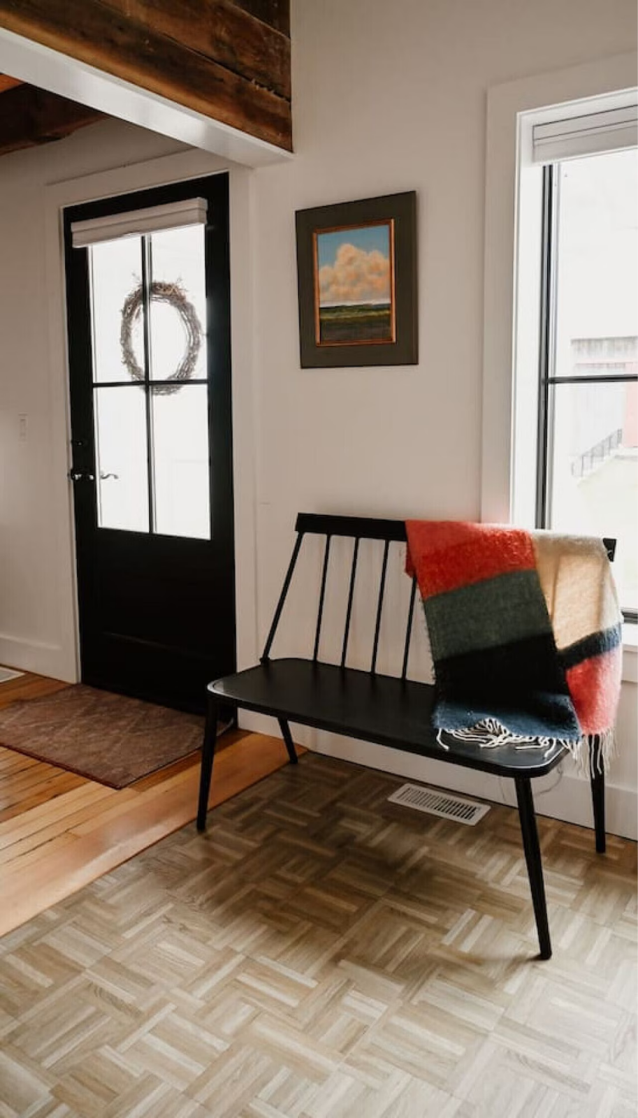 Interior view of a cozy room featuring a black door with a wreath, a black bench with a colorful blanket, a framed landscape painting on the wall, large windows, and a section of wooden and patterned flooring.