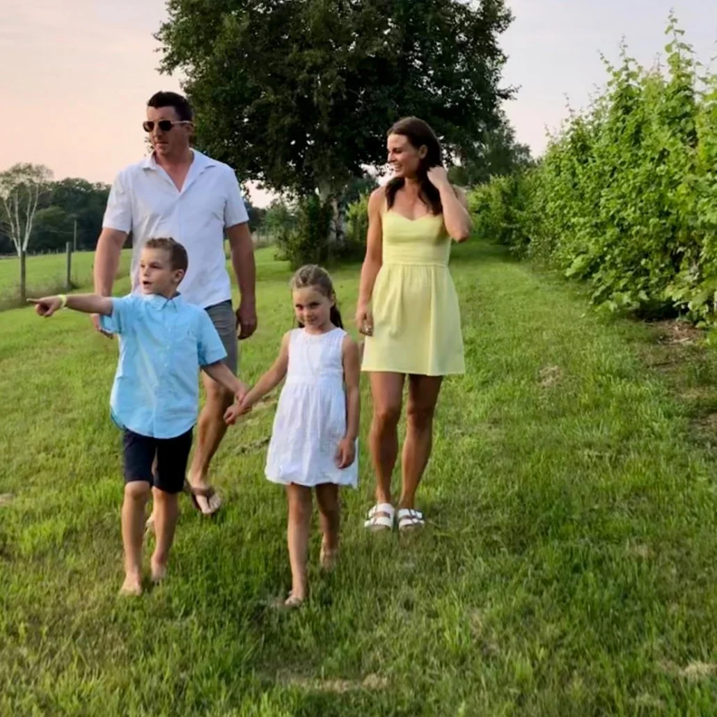 A family of five walking through a green field on a sunny day. The father is wearing sunglasses and a white shirt. The mother is in a yellow dress. The two children, a boy and a girl, are barefoot; the boy is pointing while the girl holds the mother'