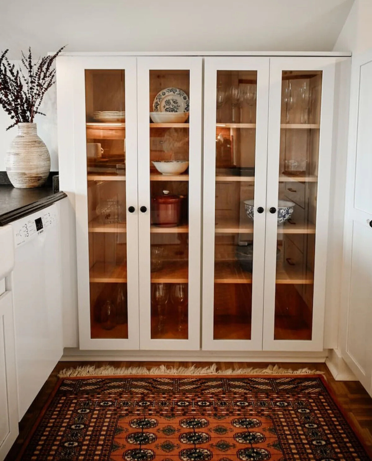 A white kitchen cabinet with glass doors, containing bowls, plates, and glasses inside. There is a decorative rug on the floor in front of the cabinet and a vase with dried flowers on the countertop to the left.
