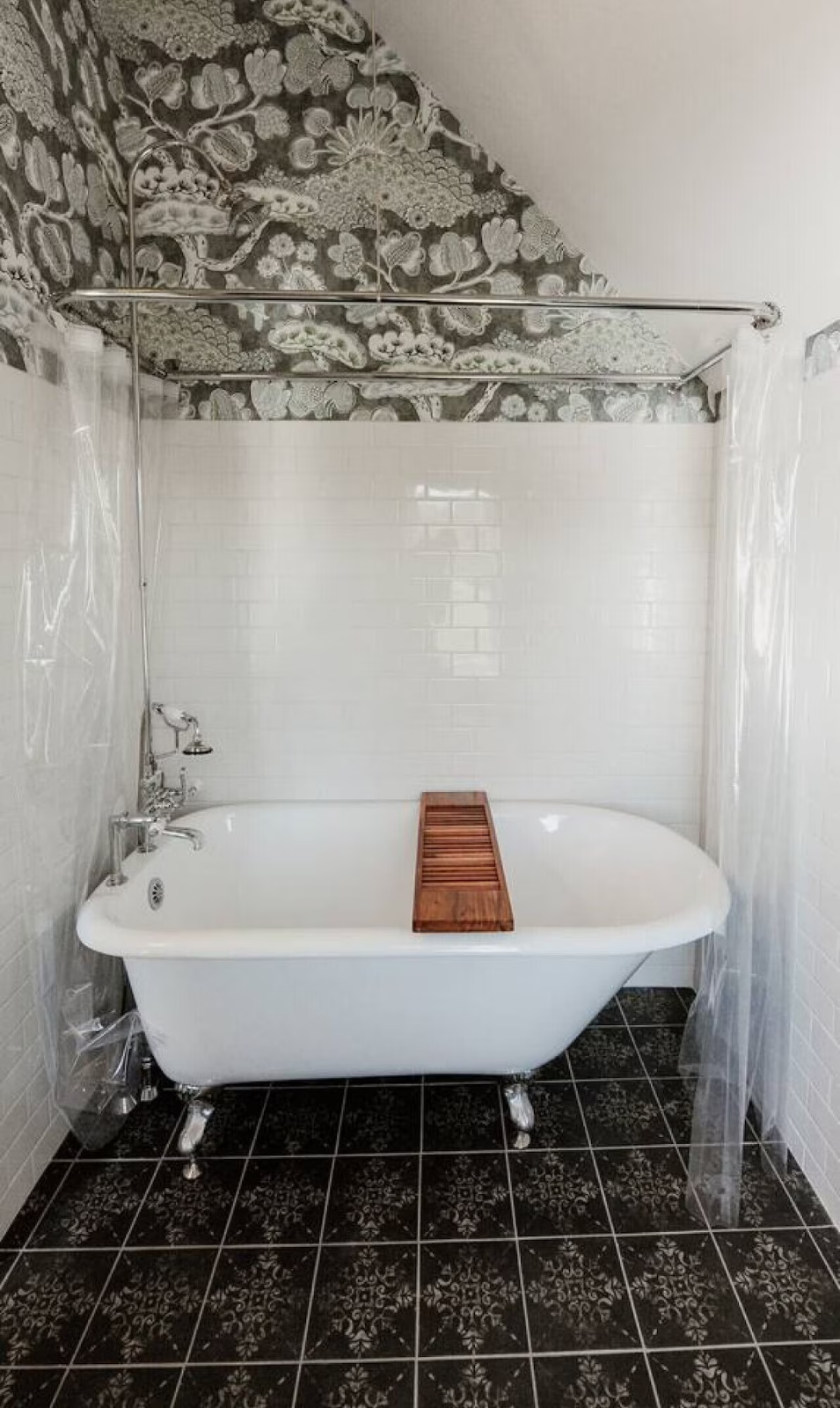 A vintage clawfoot bathtub with a wooden bath tray, in a bathroom with white subway tiles on the wall and black patterned floor tiles. The ceiling has decorative black and white floral wallpaper.