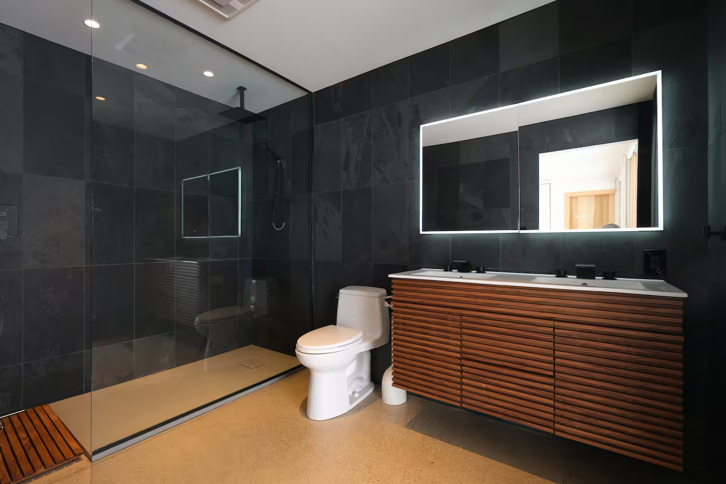 Modern bathroom with dark tiled walls, a glass-walled shower area, a white toilet, and a wooden vanity with a large mirror illuminated by LED lights.