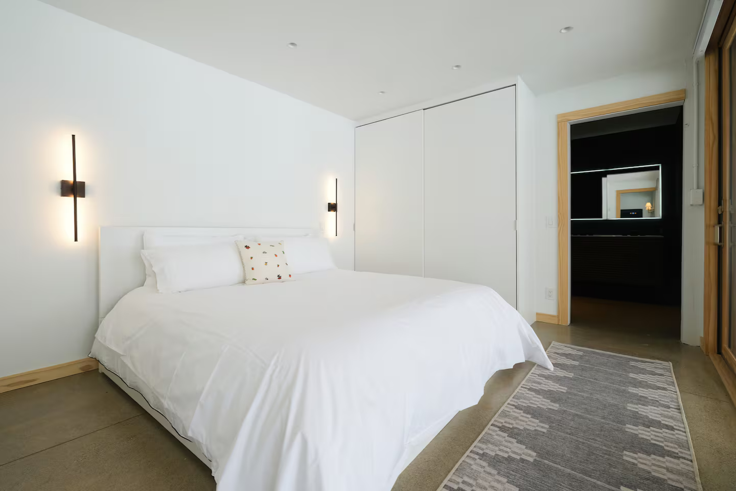 Modern bedroom with white walls, bed with white bedding, small decorative pillow, and wall-mounted lights