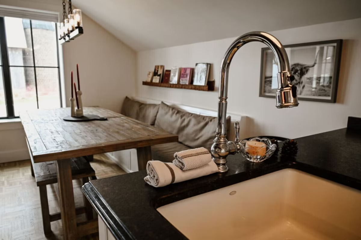 View of a kitchen with a sink and countertop in the foreground, a wooden dining table with candles, and a couch with a shelf of books on the back wall in the background.