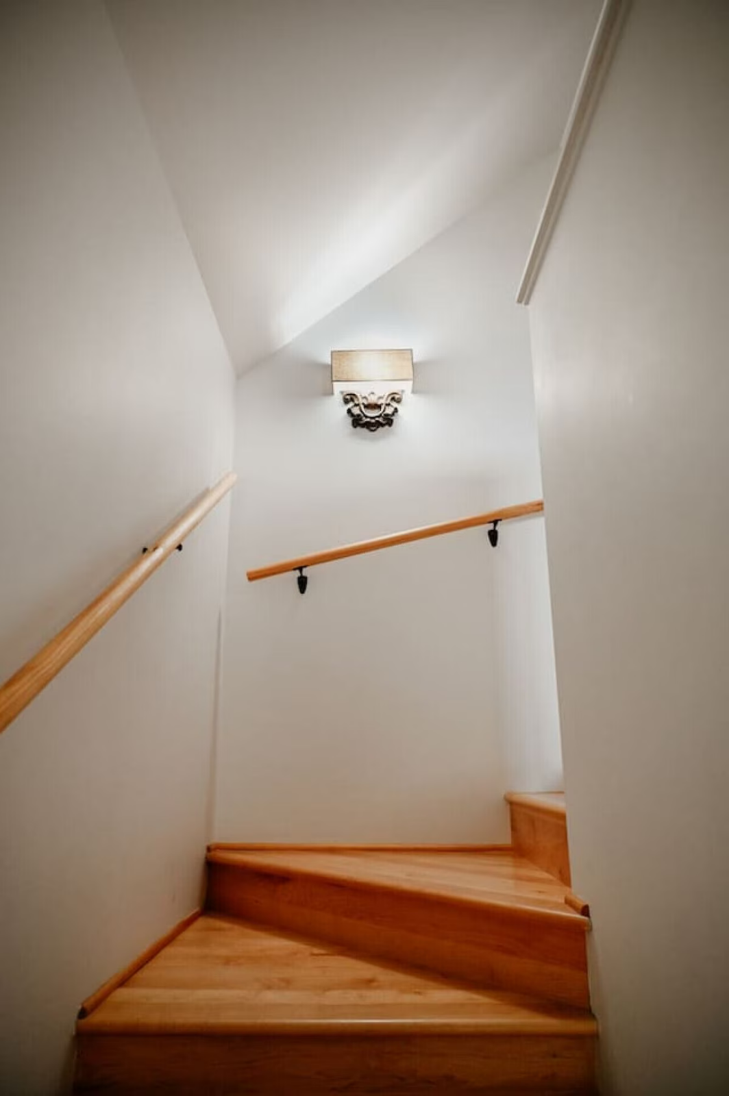Wooden staircase with beige walls, handrails on both sides, and a decorative wall-mounted light fixture at the top.