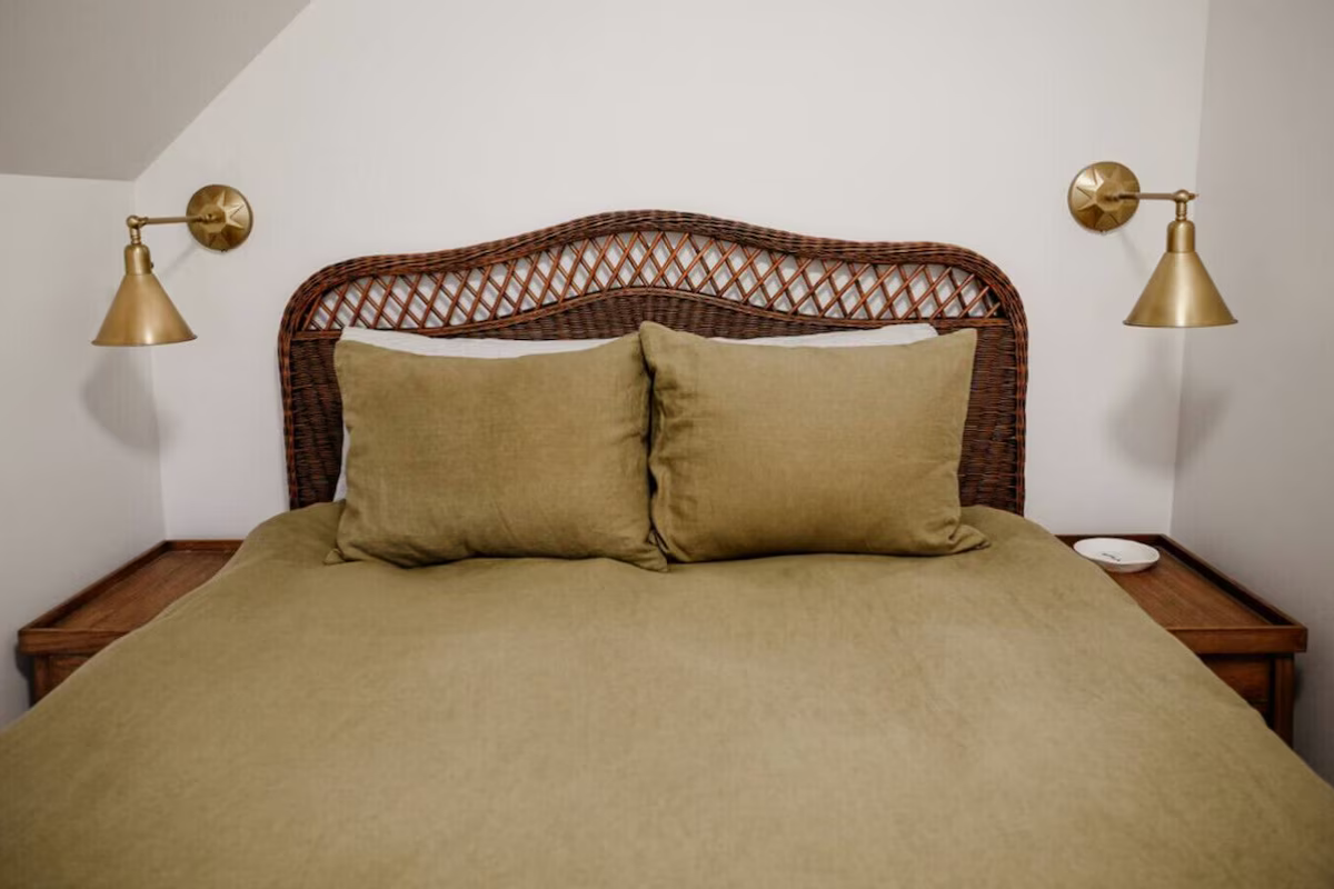 A neatly made bed with a woven headboard, greenish bedspread, two matching pillows, and two brass wall-mounted lamps on either side at night.