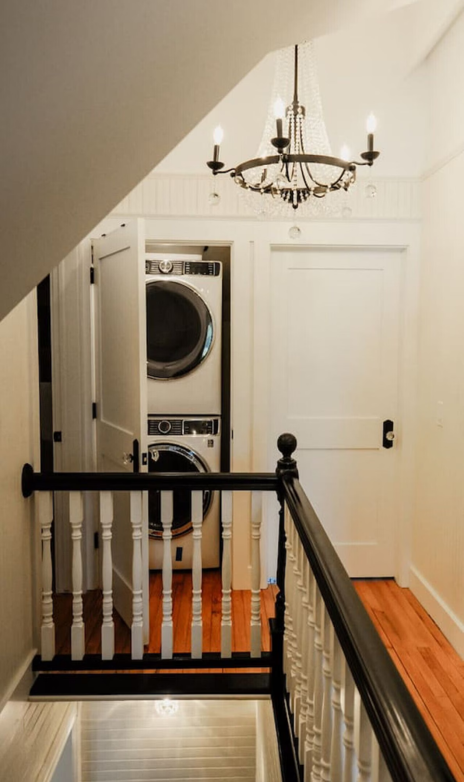 A staircase landing with a black and white laundry area in the background featuring stacked washer and dryer units and a white door. A chandelier hangs from the ceiling.