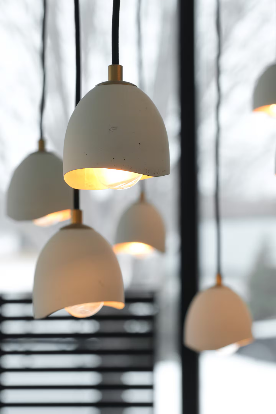 Multiple white pendant lights hanging from the ceiling with black cords, illuminated with warm yellow light, in a room with large windows and black window frames.