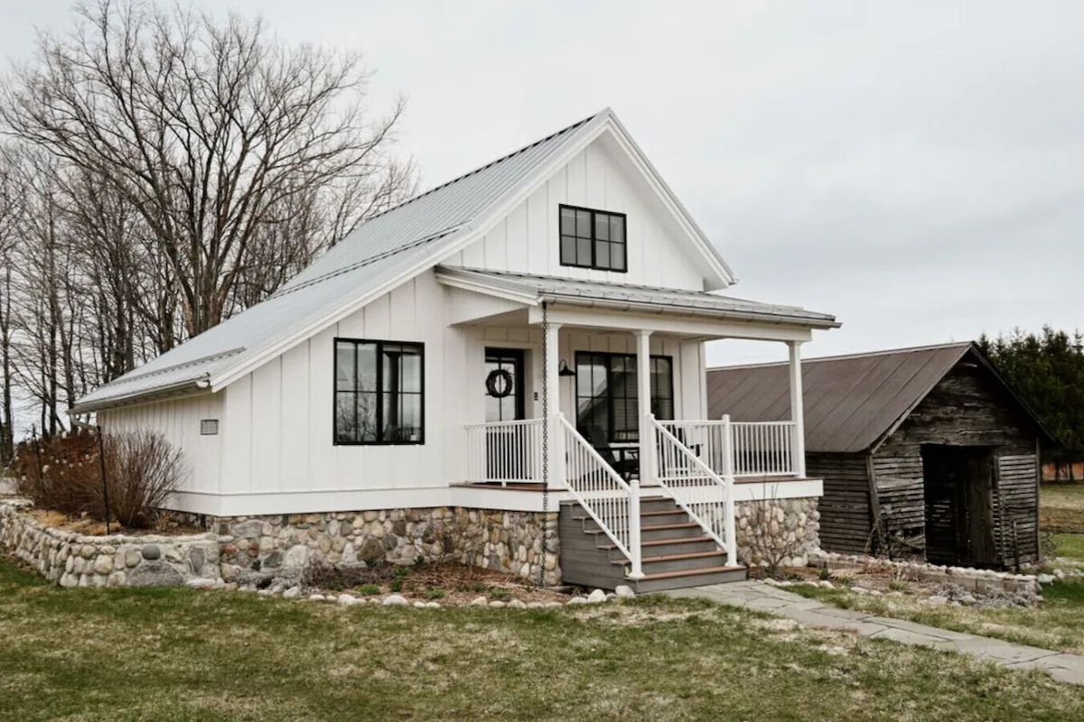 White house with black window frames and a stone foundation, elevated with stairs leading to porch, next to an old wooden shed in a yard with trees in the background.