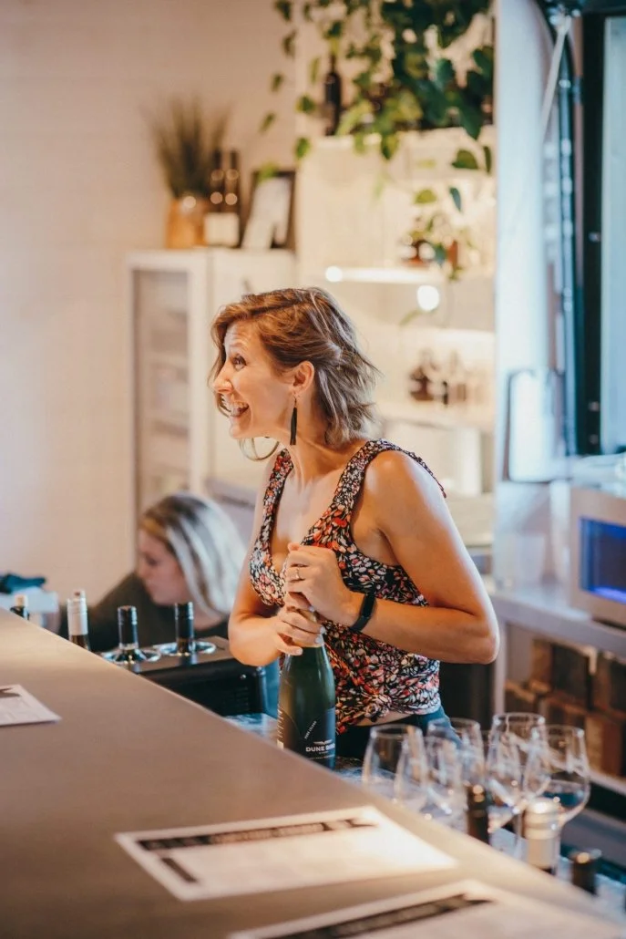 A woman with short blonde hair smiling behind a bar, holding a bottle of sparkling wine or champagne. There are wine glasses on the bar in front of her and a woman with blonde hair working in the background. The setting appears to be a cozy, well-dec