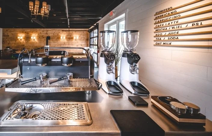 Coffee shop counter with two coffee grinders, espresso machine, sink, and menu board listing drinks like espresso, cappuccino, and latte.
