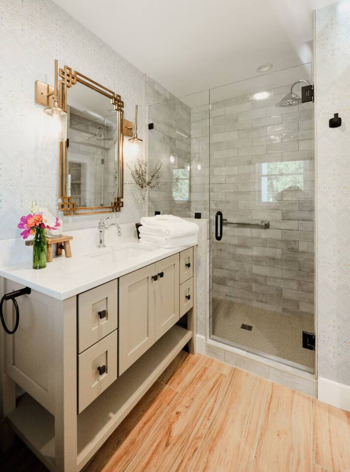 Bathroom with beige vanity, white countertop, large mirror, decorative wall lights, and a walk-in shower with glass doors and gray tiles.