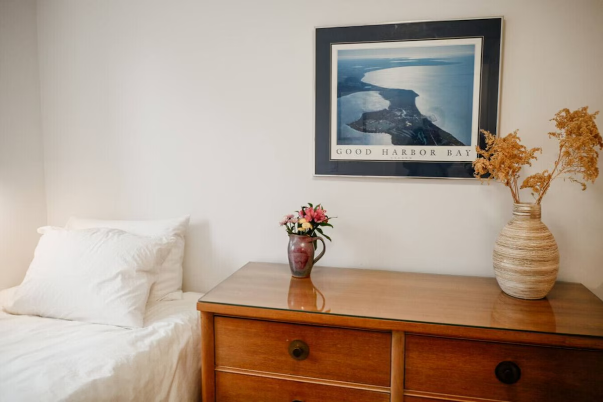 A bedroom with a bed covered in white linens on the left, a wooden dresser in the center, a framed aerial photo of Good Harbor Bay on the wall above, a small vase with pink and yellow flowers on the dresser, and a large beige vase with dried orange f