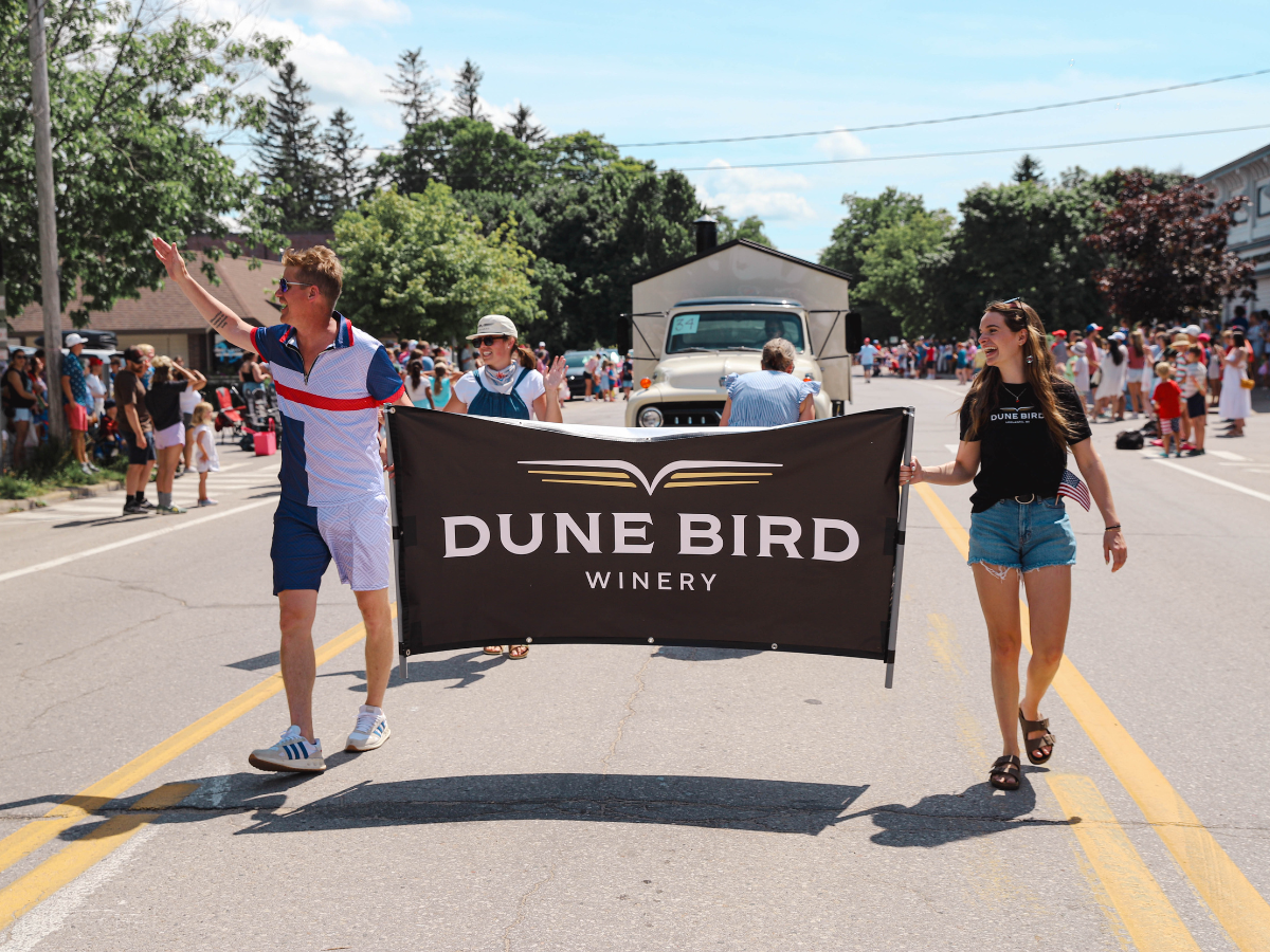 Two people holding a "Dune Bird Winery" banner at a parade with onlookers and vintage vehicles in the background on a sunny day.