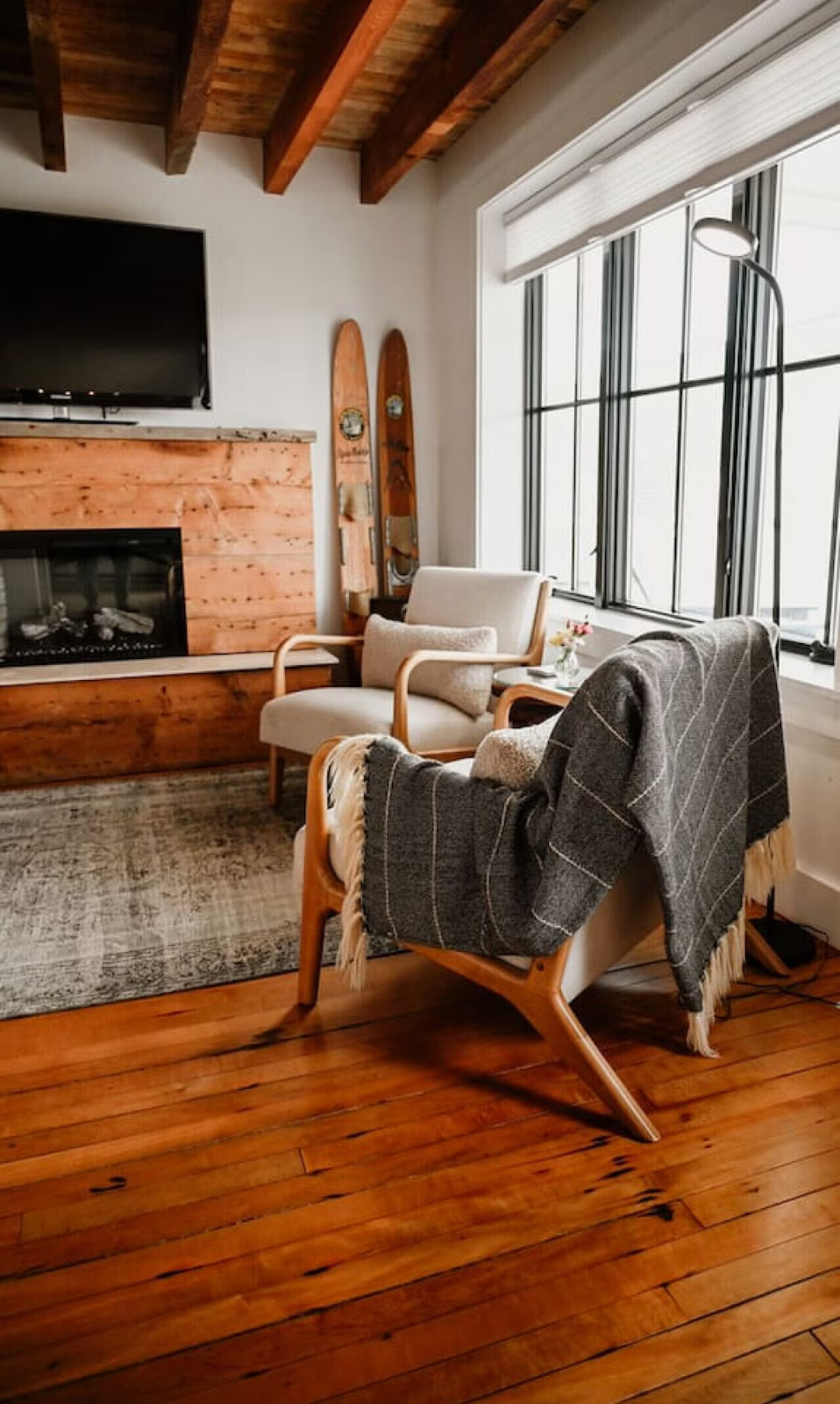 Living room with wooden floor and ceiling, fireplace with wooden mantel, flat-screen TV mounted on wall above fireplace, two vintage wooden skis leaning against wall near fireplace, beige armchair with throw pillow, gray armchair with throw blanket, 