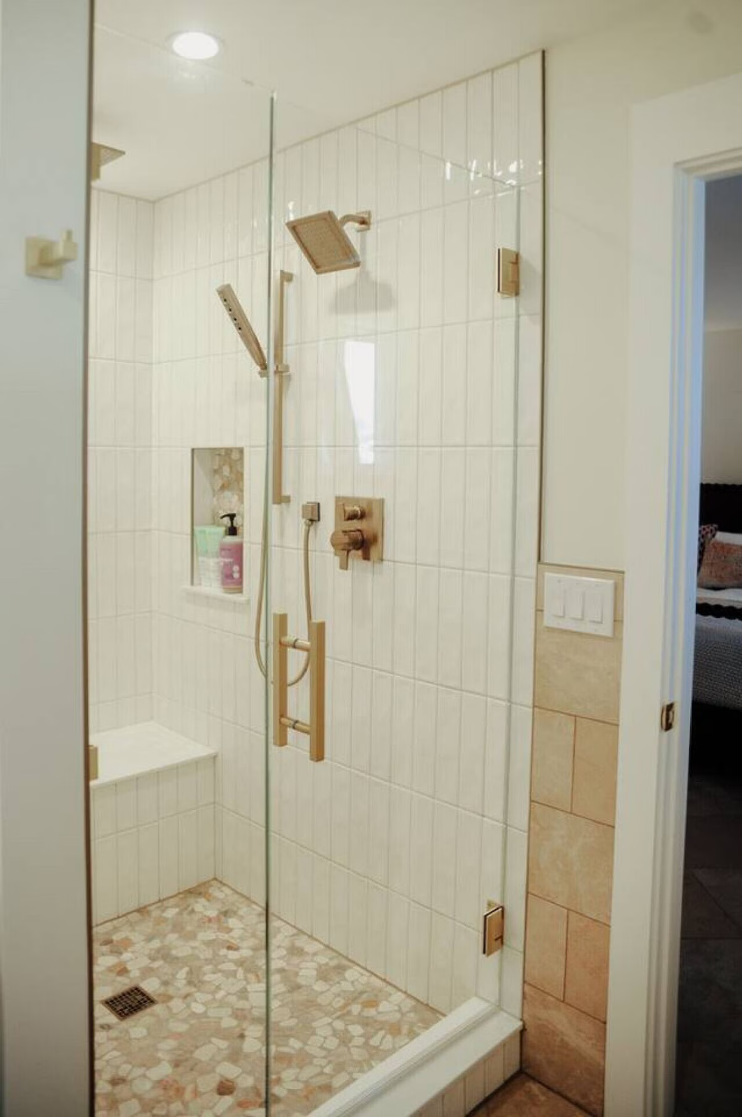 A walk-in shower with a glass door, white tiled walls, and a mosaic tile floor, featuring a bench, a built-in shelf with soap, and multiple showerheads with control fixtures.