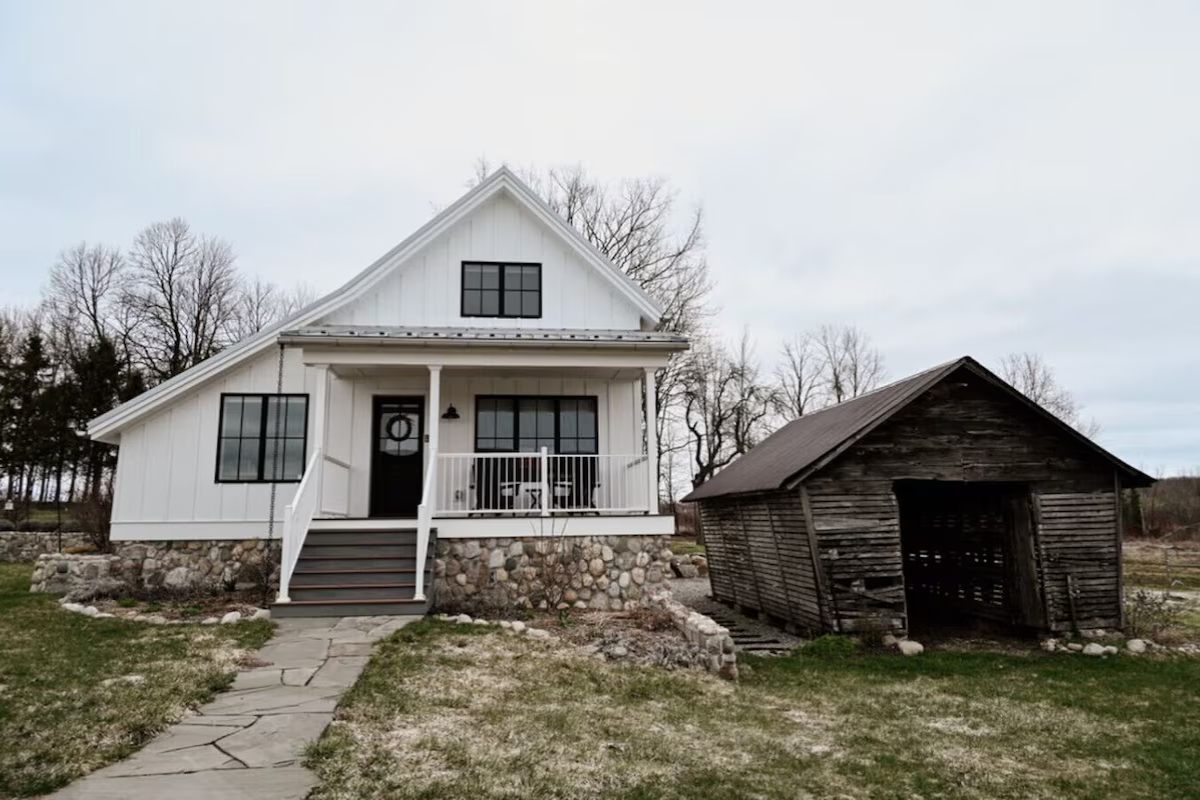 Renovated historic granary turned chic rental.  Original stone foundation, black-framed windows, and a small porch with steps; next to it is an old, weathered wooden shed. Located in Leelanau County near Lake Michigan.