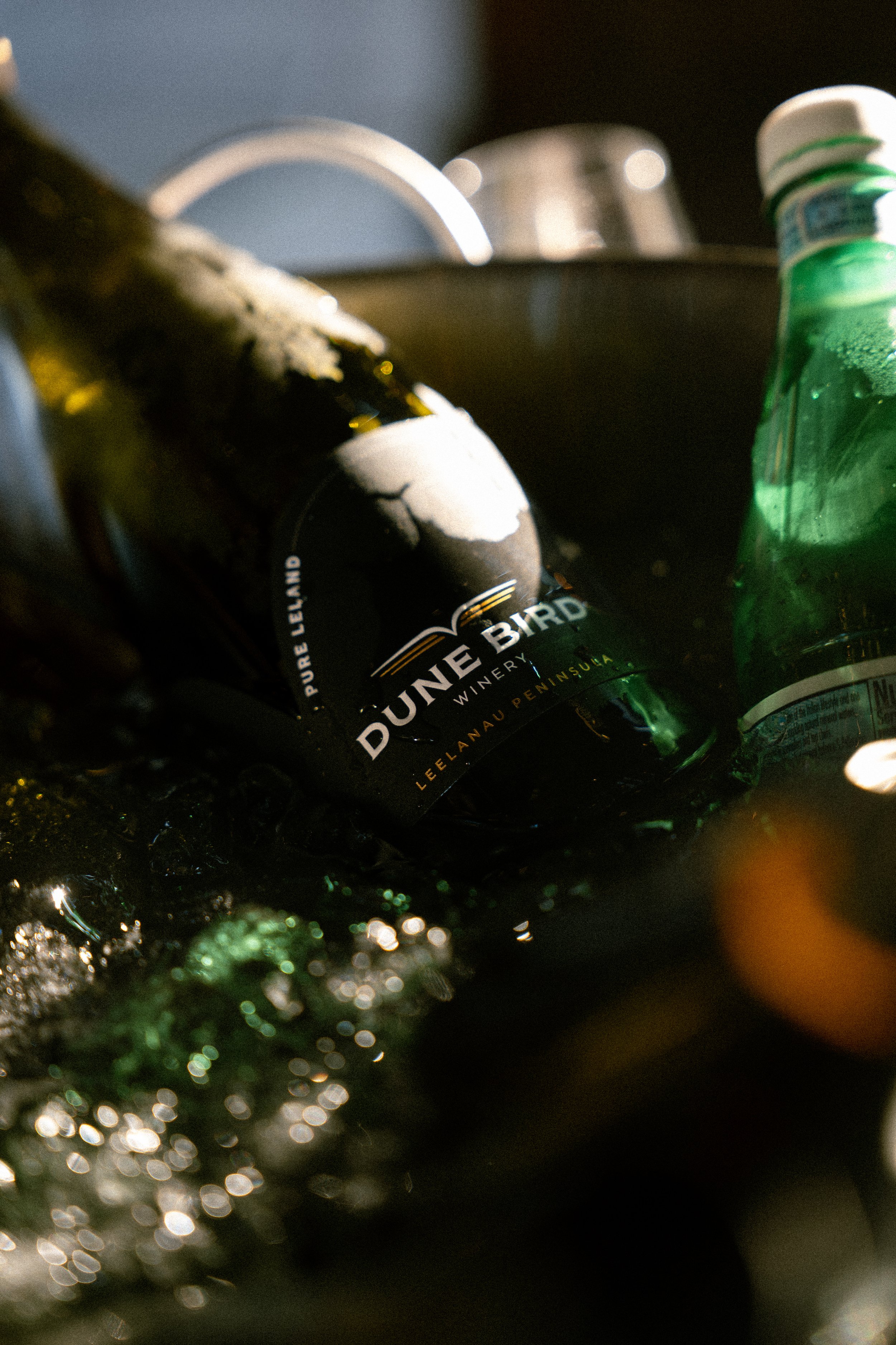 Close-up of a beer bottle labeled 'Dune Bird Winery,' lying on a wet, reflective surface with other bottles and glasses blurred in the background.