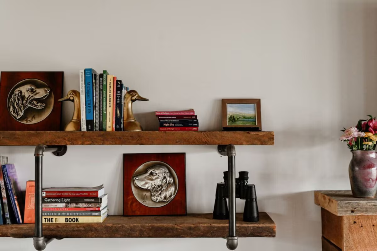 Wooden wall shelf with decorative books, brass duck figurines, framed artwork, binoculars, and a flower vase.