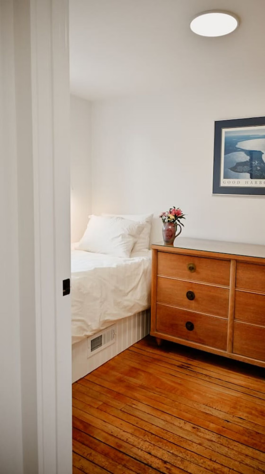 A cozy bedroom with a white bed, a wooden dresser, a decorative vase with pink flowers, and nautical-themed artwork on the wall.