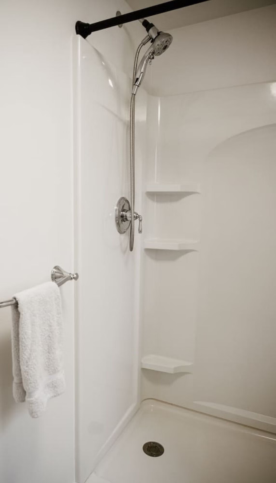 Empty white shower stall with built-in shelves, a handheld showerhead, and a towel bar with a white towel.