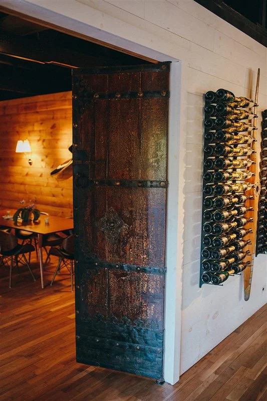 A rustic, partially open sliding door made of weathered metal on a white wall, with a wine rack holding several bottles of wine mounted on the wall beside it. A warmly lit dining area with wooden walls and chairs is visible through the doorway.