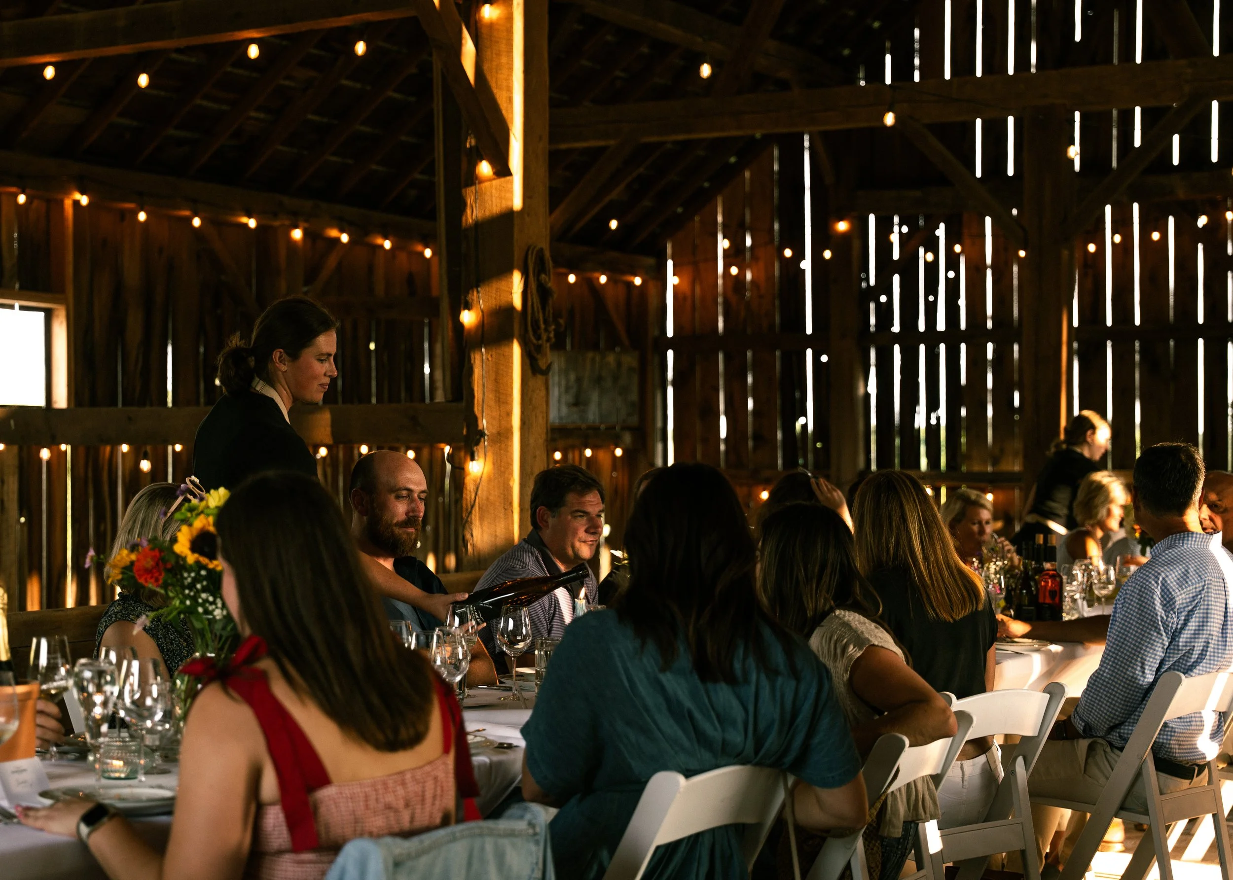 A group of people sitting at a long dinner table inside a rustic barn with wooden walls and string lights hanging from the ceiling. A woman is serving or pouring drinks, and there are flowers and wine glasses on the table.